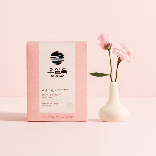 Boîte rose poudré OSULLOC Wedding Green Tea à côté d'un vase avec des fleurs de lys roses, sur un fond minimaliste.