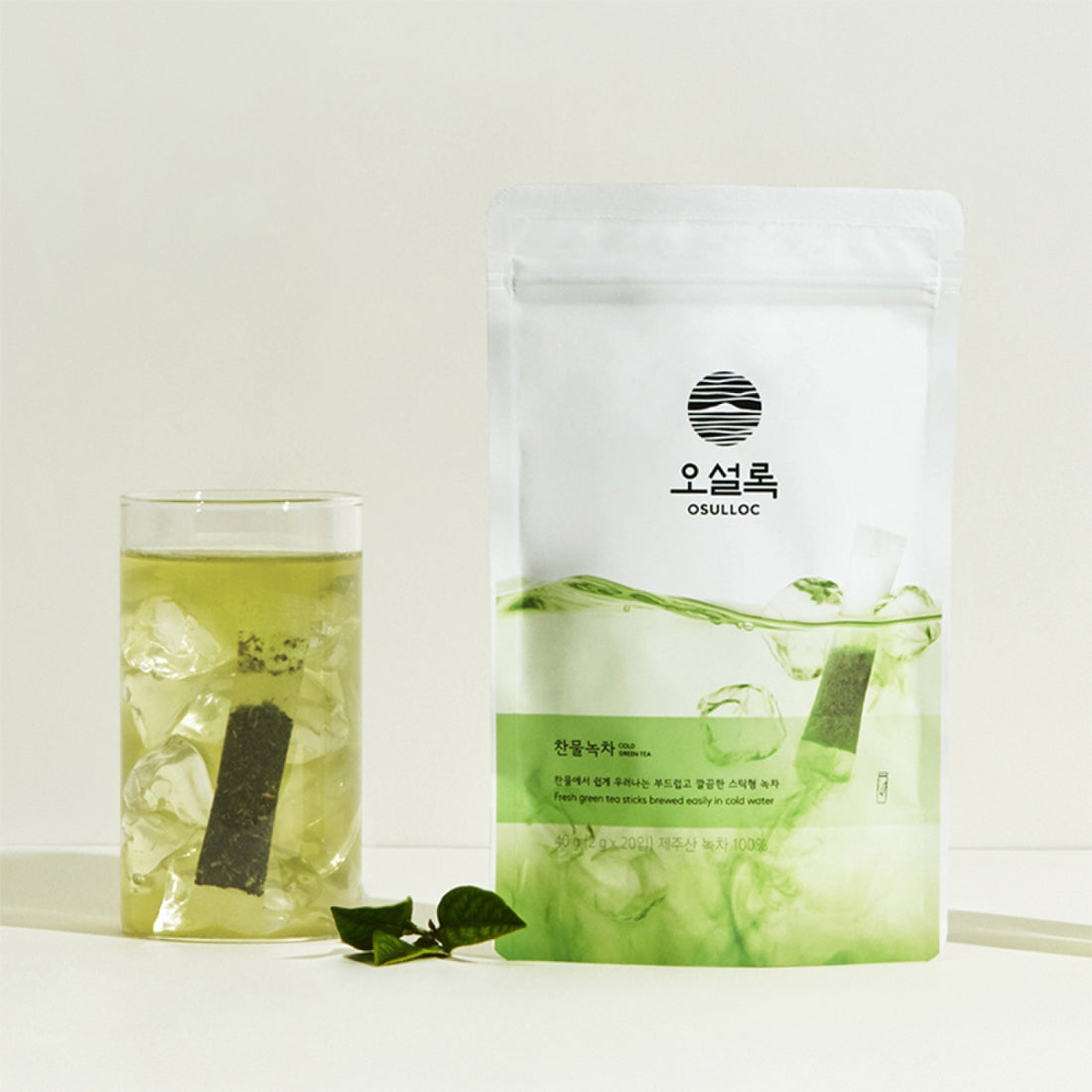 Sachet blanc OSULLOC Cold Green Tea avec un verre de thé vert glacé et des feuilles de thé fraîches.