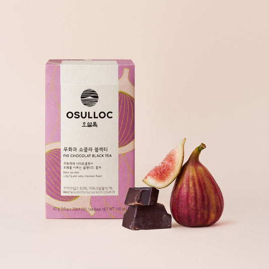 Boîte de thé OSULLOC Fig Chocolate sur un fond d'herbe avec des figues fraîches coupées et des fèves de cacao.